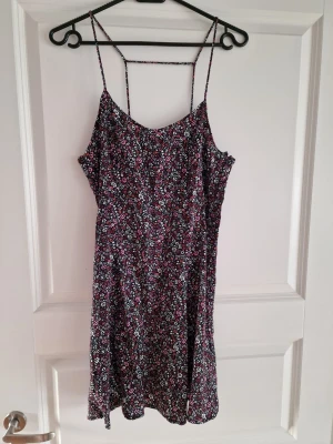 Ny blommig kort klänning med smala band med etikett - Söt kort klänning med smala axelband och blommigt mönster i svart, vitt, rosa och blått. Klänningen har en avslappnad passform och är perfekt för varma dagar. Tillverkad i ett lätt och luftigt material som känns skönt mot huden.