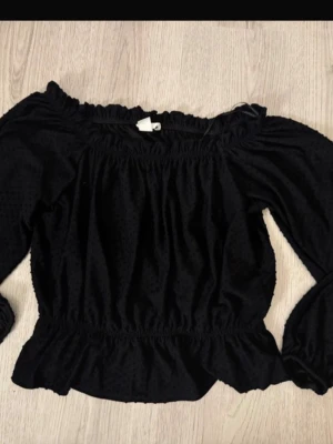 Svart offshoulder blus med volang - Snygg svart offshoulder blus med volangkant och långa ärmar. Blusen har resår vid axlar och midja för en bekväm passform och är tillverkad i ett lätt, strukturerat tyg. Perfekt för dig som vill ha en trendig och feminin look.