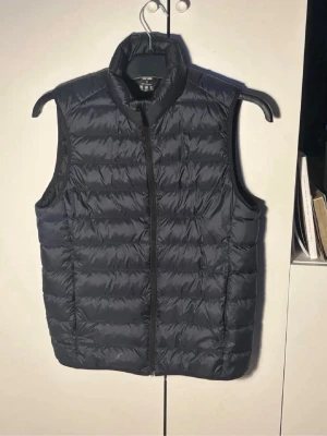 Uniqlo väst  - Snygg blå dunväst med hög krage och dragkedja framtill. Västen har en quiltad design med horisontella sömmar och är lätt vadderad för extra värme. Perfekt att slänga över en hoodie eller tröja för en chill look.