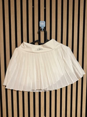 Vit tennis kjol från Alo - Snygg vit kortkjol från Alo med plisserad design och bred midjeresår. Kjolen har en sportig känsla och passar perfekt till en aktiv stil. Tillverkad i ett lätt och bekvämt syntetmaterial.
