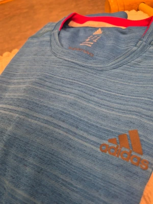 Blå långärmad träningströja Adidas L - Snygg blå långärmad träningströja från Adidas med diskret mönster och logga på bröstet. Tröjan har rund halsringning med rosa insida och dekorativa ränder i brons på sidorna. Perfekt för löpning eller gym. Tillverkad i funktionsmaterial.