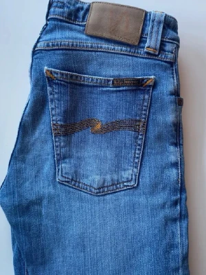 Blå jeans från Nudie Jeans ”Lean Dean” - Säljer ett par klassiska blå jeans från Nudie Jeans med smal passform och snygga kontrastsömmar. Jeansen har en bakficka med den ikoniska vågade sömmen och patch i midjan. Tillverkade i mjuk denim som sitter skönt hela dagen.