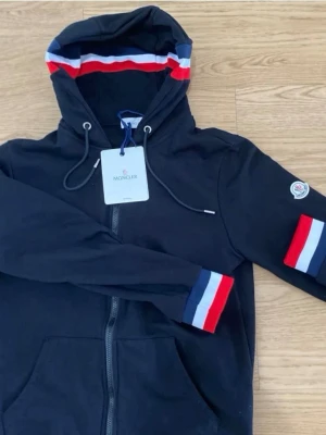 Svart Moncler hoodie  - Snygg svart hoodie från Moncler med dragkedja, huva och dragsnören. Röd, vit och blå randdetalj på ärmslut och insidan av huvan. Moncler-logga på ena ärmen. Perfekt för en stilren och sportig look.