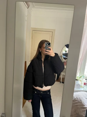 Svart cropped pufferjacka - Säljer en svart, kort pufferjacka med hög krage och dragkedja framtill. Jackan har en boxig passform och breda ärmar, perfekt för att hålla värmen under kalla dagar. Materialet är quiltat och fylligt, vilket ger en trendig look.