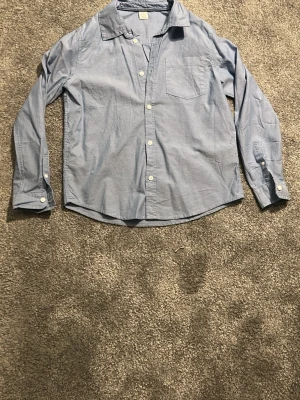 Ljusblå skjorta med ficka - En klassisk ljusblå skjorta i bomull med vit knappstängning och bröstficka. Skjortan har lång ärm och avslappnad passform, perfekt för en clean och enkel stil.