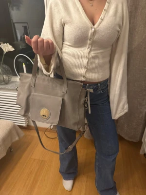 Beige handväska med guldspänne - Stilren beige handväska i rektangulär form med både handtag och axelrem. Väskan har ett stort ytterfack med lock och ett runt guldspänne framtill. Perfekt för dig som gillar en klassisk och enkel look. Materialet ser ut att vara konstläder.