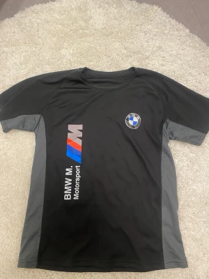 BMW M Motorsport svart t-shirt - Snygg svart t-shirt från BMW M Motorsport med logga och tryck i vitt, blått och rött på framsidan. T-shirten har grå paneler på sidorna och är tillverkad i ett sportigt, lätt material som passar perfekt för aktiva dagar.