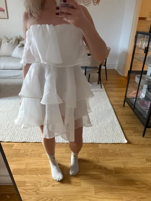 Vit volangklänning axelbandslös - Säljer en superfin vit kortklänning med flera lager volanger och axelbandslös design. Klänningen har öppen rygg och är perfekt för sommarens alla tillfällen. Materialet känns lätt och luftigt.