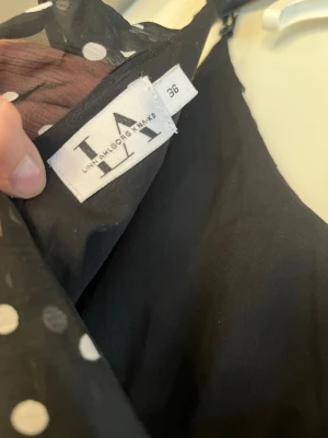 Svart prickig volangklänning Lina Ahlborg - Supersöt svart kortklänning från Lina Ahlborg x NA-KD med vita prickar och flera lager volanger. Klänningen har smala axelband, v-ringning och luftig passform. Perfekt för dig som vill ha en lekfull och trendig look.