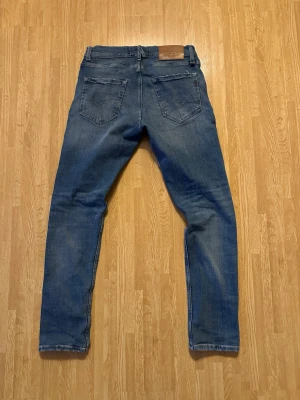 Blå slitna jeans från Replay - Säljer ett par blå jeans från Replay med slim fit och slitningar på framsidan för en trendig look. Jeansen har klassiska fem fickor, knappgylf och läderpatch bak i midjan. Perfekta för dig som gillar en avslappnad och modern stil.