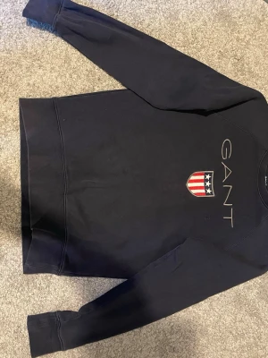 Svart sweatshirt från GANT med logga - Snygg svart sweatshirt från GANT med broderad logga och sköld med stjärnor och ränder på bröstet. Klassisk rund halsringning och långa ärmar. Tillverkad i mjukt bomullsmaterial för en bekväm känsla.