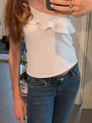 Vit oneshoulder topp med volang - Snygg vit oneshoulder topp med bred volangdetalj över axeln. Perfekt till jeans eller kjol för en trendig look. Tillverkad i mjukt bomullsmaterial som känns skönt mot huden. Mango teen topp storlek M men mer som S/xs