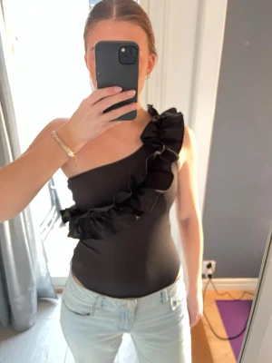 Svart oneshoulder topp med volang - Säljer en svart oneshoulder topp med stor volang längs halsringningen. Lappen är borta men köpt på Sellpy 