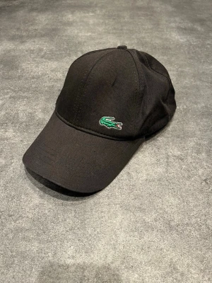 Svart keps från Lacoste - Svart keps från Lacoste med klassisk böjd skärm och broderad grön krokodil på framsidan. Baktill finns vit brodyr med Lacoste-loggan. Kepsen är tillverkad i bomull och har en stilren och sportig look.