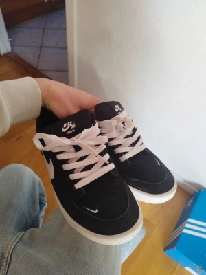 Svarta Nike SB sneakers med vit sula - Säljer ett par svarta Nike SB sneakers med vit sula och vita skosnören. Skorna har klassisk låg profil, tydlig Nike-logga på sidan och tungan, samt är tillverkade i mockaliknande material. Perfekta för dig som gillar streetwear och skate. Priset kan diskuteras! 😁