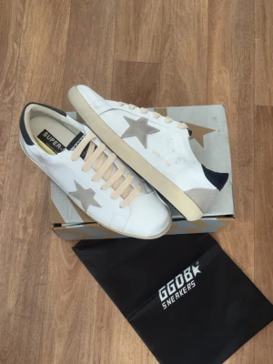 Golden Goose vita sneakers med stjärna - Golden Goose sneakers i vitt läder med grå mockadetaljer och ikonisk stjärna på sidan. Beige snörning, mörkblå häl och vintage-inspirerad sula. Klassisk låg modell med GGDB-logga på sidan och plös.Hör av er vid intresse