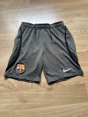 Barça shorts från Nike, grön - Gröna FC Barcelona shorts från Nike med svart detalj längs sidorna och klubbmärke på vänster ben. Tillverkade i lätt och ventilerande material, perfekta för träning eller match. Elastisk midja med dragsko för skön passform och två sidofickor. Storleken är 137-147.
