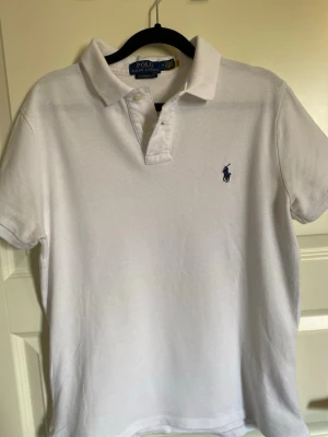 Vit piké från Polo Ralph Lauren - Klassisk vit pikétröja från Polo Ralph Lauren med korta ärmar och blå logga broderad på bröstet. Tröjan har krage, knappar vid halsen och är i en slim fit-modell. Tillverkad i mjuk bomull som känns skön mot huden.