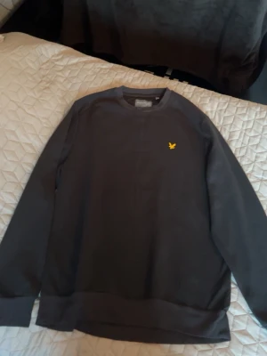 Mörkblå sweatshirt från Lyle & Scott - Snygg mörkblå sweatshirt från Lyle & Scott med klassisk rund hals och gul logga broderad på bröstet. Tröjan har långa ärmar och ribbade muddar vid ärmslut och nederkant. Perfekt för en avslappnad stil. storlek L men passar också M