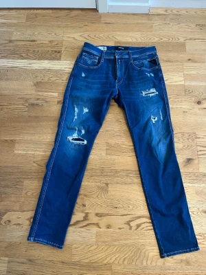 Blå slitna jeans från Replay - Säljer ett par blå jeans från Replay med slitningar och hål på framsida. Jeansen har klassisk femficksmodell, raka ben och tydliga slitdetaljer för en trendig look. Materialet är denim med stretch för extra komfort. Det är slim fit och storleken W32 L32. Bara att höra av sig vid frågor