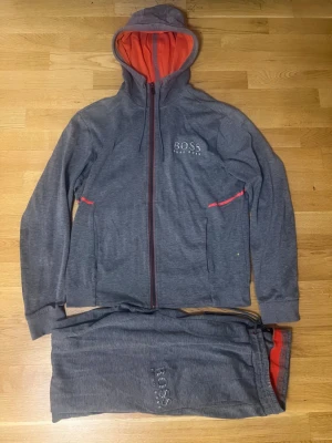 Hugo boss dress - Mysigt loungewear-set från BOSS i grått med röda detaljer. Setet består av en zip-hoodie med huva och matchande byxor, båda med BOSS-logga framtill. Mjukt material och bekväm passform, perfekt för chill dagar hemma.
