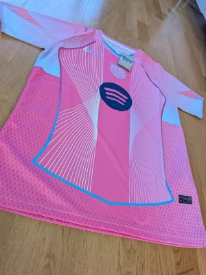 Rosa FC Barcelona matchtröja Nike - Säljer en rosa och vit FC Barcelona matchtröja från Nike med grafiskt mönster och blå detaljer. Tröjan har klubbmärke och sponsorlogga på bröstet samt korta ärmar. Perfekt för dig som älskar fotboll och vill sticka ut på planen.