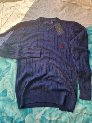 Mörkblå kabelstickad tröja Polo Ralph Lauren - Kabelstickad mörkblå tröja från Polo Ralph Lauren med rund halsringning och röd broderad logga på bröstet. Tröjan har långa ärmar och ribbade muddar. Perfekt för lager-på-lager och klassisk stil.