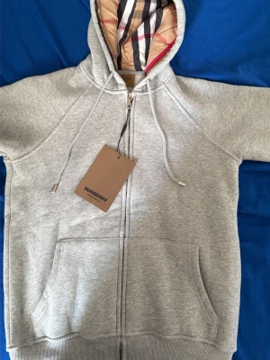 Grå zip hoodie från Burberry - Snygg grå zip hoodie från Burberry med klassiskt rutigt foder i huvan. Hoodien har dragsko i huvan, två stora fickor framtill och ribbade muddar. Tillverkad i mjuk bomullsblandning och har en exklusiv känsla. Perfekt för dig som vill ha en stilren och lyxig look.