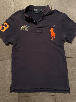 Mörkblå pikétröja Polo Ralph Lauren - Mörkblå pikétröja från Polo Ralph Lauren i slim fit med orange broderad logga och siffra på ärmen. Klassisk krage och korta ärmar, tillverkad i bomull. Snygga broderade detaljer på bröstet för en sportig look. Tröjan är något solblekt och har mindre fläckar under armarna 