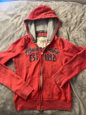Röd hoodie från Abercrombie & Fitch - Röd hoodie med dragkedja från Abercrombie & Fitch. Broderad text och siffror i blått framtill, kängurufickor och fodrad huva med grå insida. Perfekt för en avslappnad och cool stil.