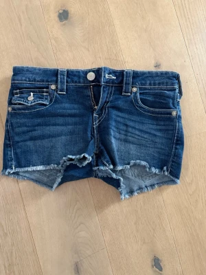 Blå jeansshorts från True Religion - Snygga blå jeansshorts från True Religion med råa kanter och broderade bakfickor. Klassisk femficksmodell med vit kontrastsöm och knappstängning. Perfekta för en avslappnad stil.