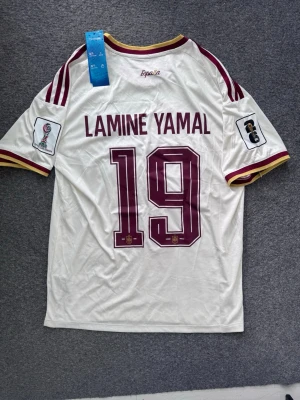 Spanien Lamine Yamal VM matchtröja - Säljer en vit Spanien fotbollströja från Adidas med Lamine Yamal och nummer 19 på ryggen. Tröjan har vinröda och gula detaljer, broderad landslagssköld och Adidas-logga framtill samt officiella turneringsmärken på ärmarna. Perfekt för fotbollsfans! OBS: ADIDAS PLASTPÅSE INGÅR I KÖPET! PRISET KAN DISKUTERAS