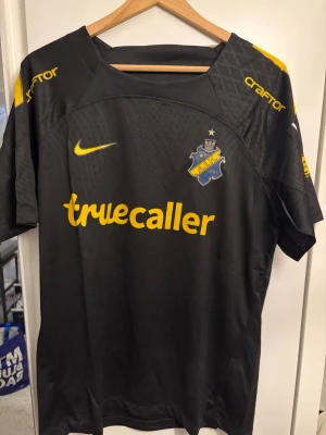 AIK matchtröja svart Nike - Säljer en svart AIK matchtröja från Nike med gula detaljer och tryck. Tröjan har AIK:s klubbmärke på bröstet, sponsorloggor som Truecaller och SVEA samt gula inslag på axlarna. Tillverkad i lätt och ventilerande material, perfekt för fotboll eller träning.