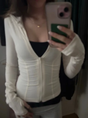 Vit tränings zip tröja - Slimmad vit träningströja  med dragkedja framtill. Långa ärmar med tumhål och markerade sömmar ger en sportig vibe. Perfekt att slänga på sig över ett linne. Materialet ser ut att vara mjukt och stretchigt, troligen bomull eller bomullsblandning.