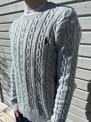 Ljusgrå kabelstickad tröja Polo Ralph Lauren - Snygg ljusgrå kabelstickad tröja från Polo Ralph Lauren med klassisk rund halsringning och broderad logga på bröstet. Tröjan har långa ärmar och ribbade muddar. Perfekt för lager-på-lager och passar till jeans eller chinos.