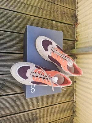 Ecco sneakers i beige och orange - Snygga sneakers från Ecco i beige mocka med orange och vinröda meshdetaljer. Skorna har klassisk låg profil, vita sulor och beige snörning. Perfekta för dig som gillar en sportig och trendig look med färgkombinationer som sticker ut.