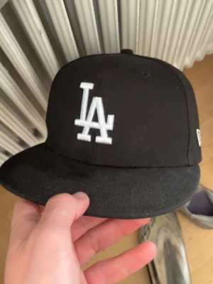 Svart LA-keps från New Era - Säljer en svart keps från New Era med LA-logga broderad i vitt framtill och MLB-logga baktill. Klassisk flat brim och stilren design som passar perfekt till streetwear. Materialet är bomull och kepsen har en strukturerad form.