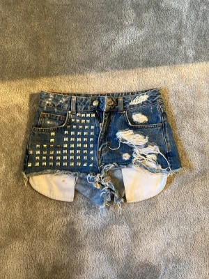Jeansshorts från Bik Bok - Snygga blå jeansshorts från Bik Bok med råa kanter, slitningar och fransar. Klassisk femficksmodell med knapp och dragkedja. Perfekta för sommaren och festivaler. Är väldigt använda och har några defekter men går defenitivt att använda!