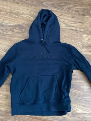 Marinblå hoodie från J.Lindeberg - Snygg marinblå hoodie från J.Lindeberg med logga på bröstet. Klassisk modell med stor huva och känguruficka framtill. Perfekt för en chill och stilren look. Mjuk bomullskänsla och ribbade muddar vid ärmslut och nederkant.