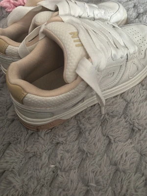 Vita Nike sneakers med beige detaljer - Snygga vita Nike sneakers med beige detaljer och breda vita skosnören. Skorna har perforerad tå för ventilation och en låg silhuett. Tillverkade i läder med mjuk vadderad insida för extra komfort. Diskret Nike-logga på sidan och tungan.
