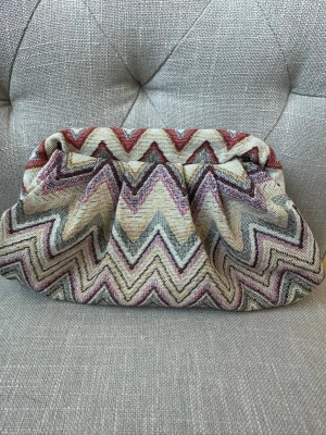 Clutchväska med zigzag-mönster - Snygg clutchväska i tyg med färgglatt zigzag-mönster. Väskan har en mjuk, puffig form och svart tygfoder på insidan. Perfekt accessoar för att lyfta din outfit. Väskan är handgjord 
