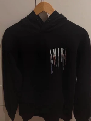 Svart amiri hoodie - Svart hoodie från Amiri med färgstarkt droppande logotyptryck både fram och bak. Klassisk modell med huva och dragsko, samt ribbade muddar vid ärmslut och nederkant. Perfekt för dig som gillar streetwear och vill sticka ut med en snygg detalj. Storlek M bra skick inga defekter!
