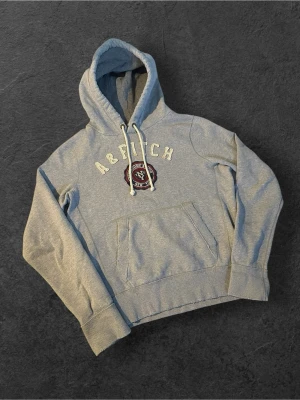 Grå hoodie från Abercrombie & Fitch | L - Säljer en grå hoodie från Abercrombie & Fitch med broderad logga och text på bröstet. Hoodien har en stor känguruficka, vita dragsnören och ribbade muddar vid ärmar och midja. Perfekt för en avslappnad och stilren look.