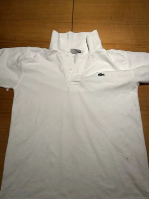 Vit polotopp från Lacoste - Vit polotopp från Lacoste med klassisk krage och knappar framtill. Toppens material är bomull och den har korta ärmar. Den ikoniska broderade krokodilen syns på bröstet. Perfekt för dig som gillar stilrena och sportiga plagg.