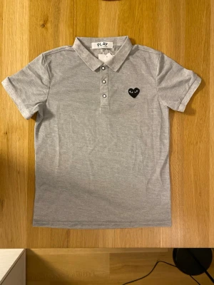 Grå pikétröja från Comme des Garçons - Snygg grå pikétröja från Comme des Garçons PLAY med klassisk krage och tre knappar framtill. På bröstet sitter det ikoniska svarta hjärta-märket med ögon. Tillverkad i mjuk bomull och har en normal passform med korta ärmar.
