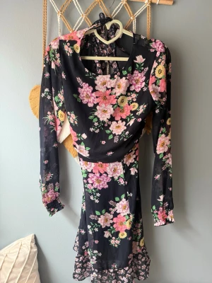 Svart blommig långärmad klänning - Svart klänning med färgglada blommor i rosa, gult och grönt. Långärmad modell med volangdetaljer och öppen rygg. Klänningen har en feminin passform och är perfekt för dig som gillar romantiska mönster. Materialet känns lätt och flowy.