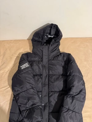 Svart pufferjacka från Burberry - Snygg svart pufferjacka från Burberry med huva och dragkedja samt tryckknappar framtill. Jackan har två fickor med dragkedja och en patch på ärmen med texten 'BURBERRY LONDON ENGLAND'. Perfekt för kalla vinterdagar.