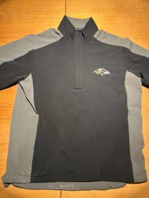 Baltimore Ravens svart/grå tröja - Cool svart och grå långärmad tröja med Ravens-logga på bröstet. Tröjan har halv dragkedja och hög krage, perfekt för att visa support för laget. Materialet är syntetiskt och känns sportigt och lätt. Passar dig som gillar amerikansk fotboll och vill ha en casual look.