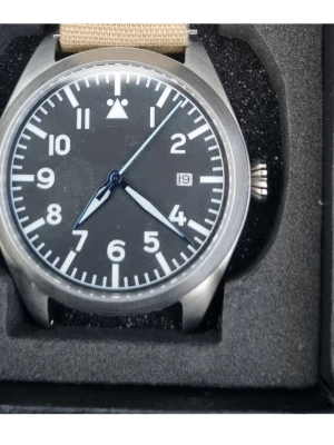 Pilotklocka med svart urtavla, typ A, Vikingwatches - Stilren pilotklocka med svart urtavla, vita siffror och index samt datumvisning. Boetten är i borstat stål och klockan har blå visare. Beige textilarmband ger en avslappnad känsla. Perfekt accessoar för dig som gillar klassisk och funktionell design från Vikingwatches.