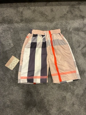 Randiga shorts med snörning - Säljer ett par snygga shorts med breda ränder i beige, svart, vit och orange. Shortsen har elastisk midja med vit snörning och sidofickor. Perfekta för sommaren och har en avslappnad passform.